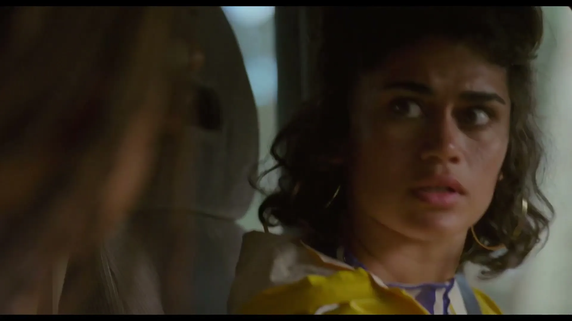 A young woman with dark curly hair and hoop earrings looks surprised while sitting in a car.