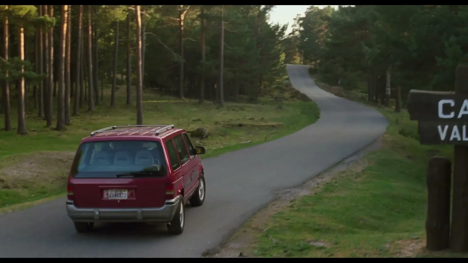 A red minivan drives away on a winding road through a forest, passing a wooden sign for a campsite.