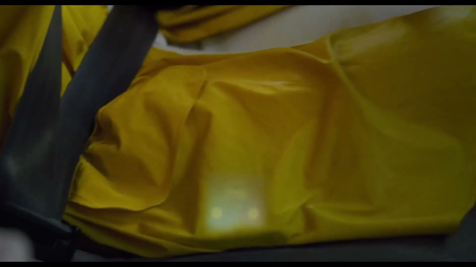 A person in a yellow raincoat is seen from a medium shot, with a seatbelt across their chest, as light reflects off the fabric.