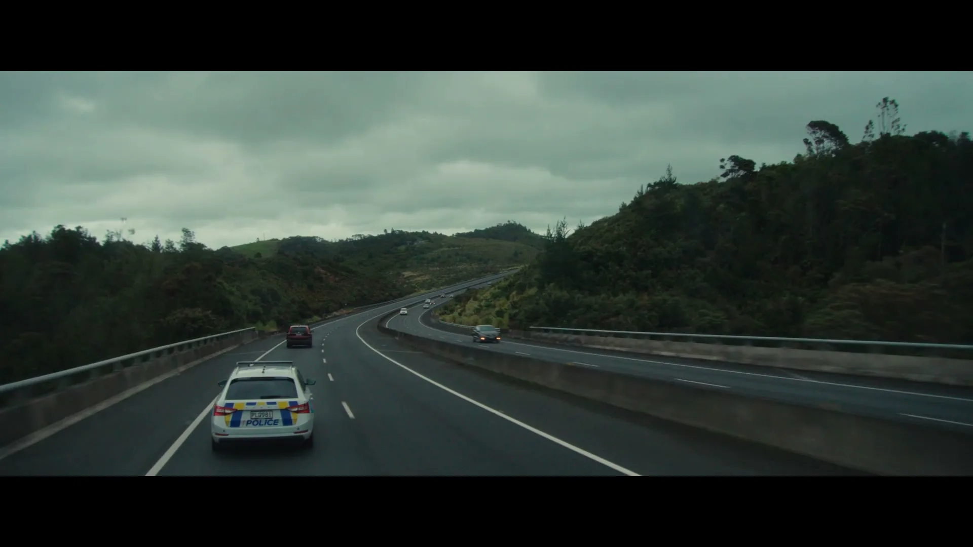 A police car drives on a multi-lane highway, followed by other cars, under a cloudy sky with green hills in the background.