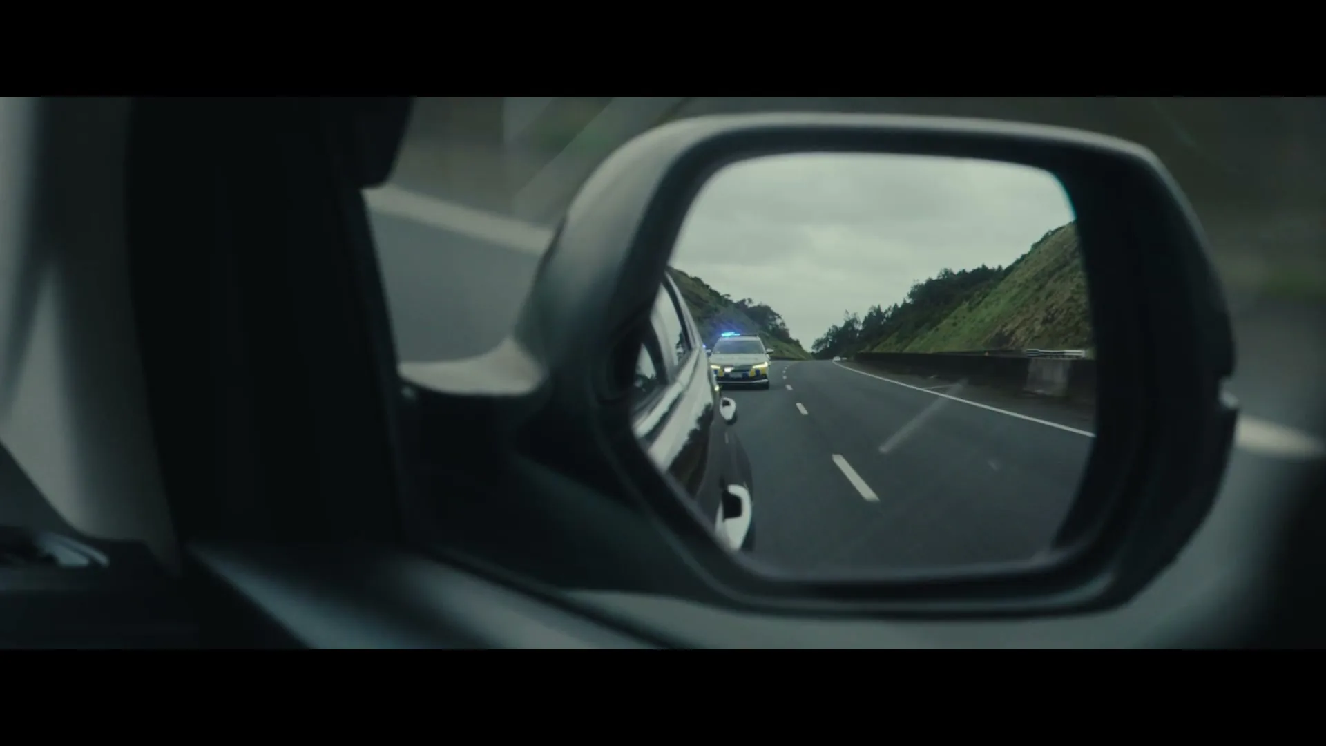 A police car with flashing lights is seen in the rearview mirror of a car driving on a highway during the day.