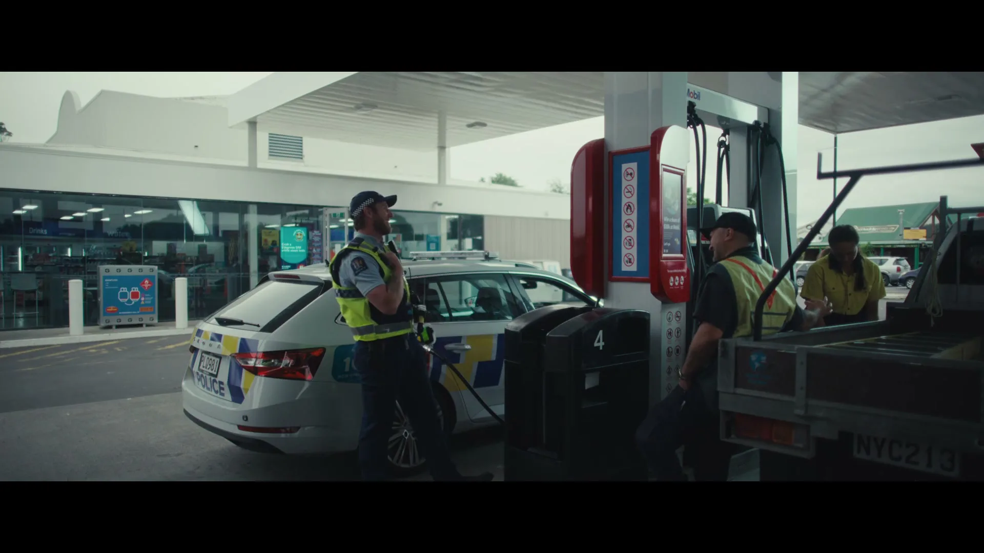 A police officer in uniform stands next to a police car at a gas station, talking to two men in work clothes.
