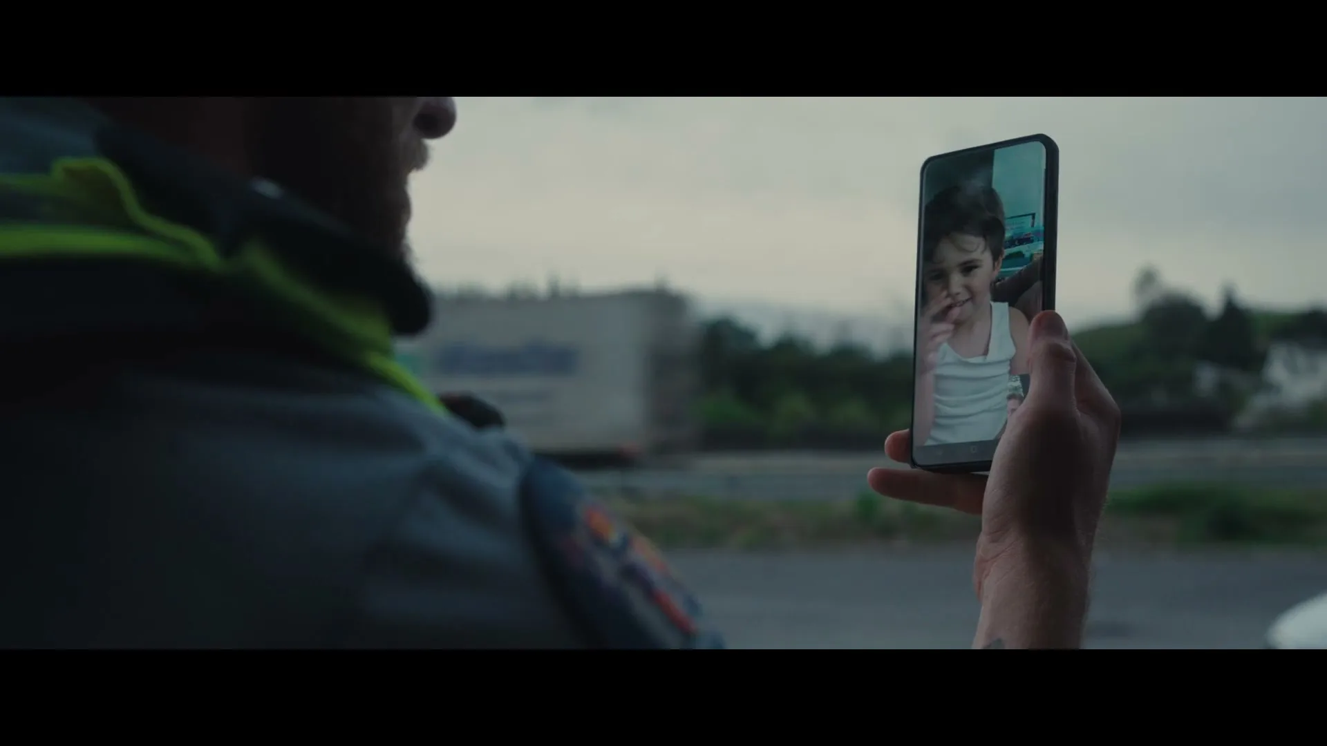 A man in uniform holds up a phone showing a video call with a smiling child, with a highway and truck in the background.