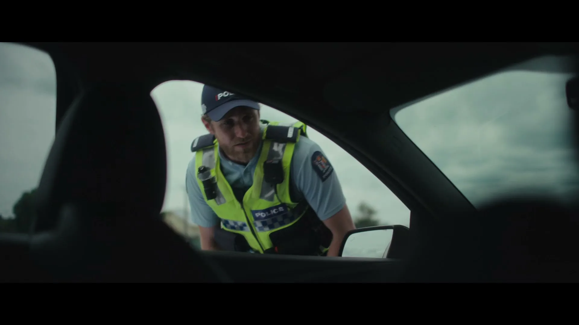 A male police officer in uniform leans down to speak to a driver through their car window on a cloudy day.