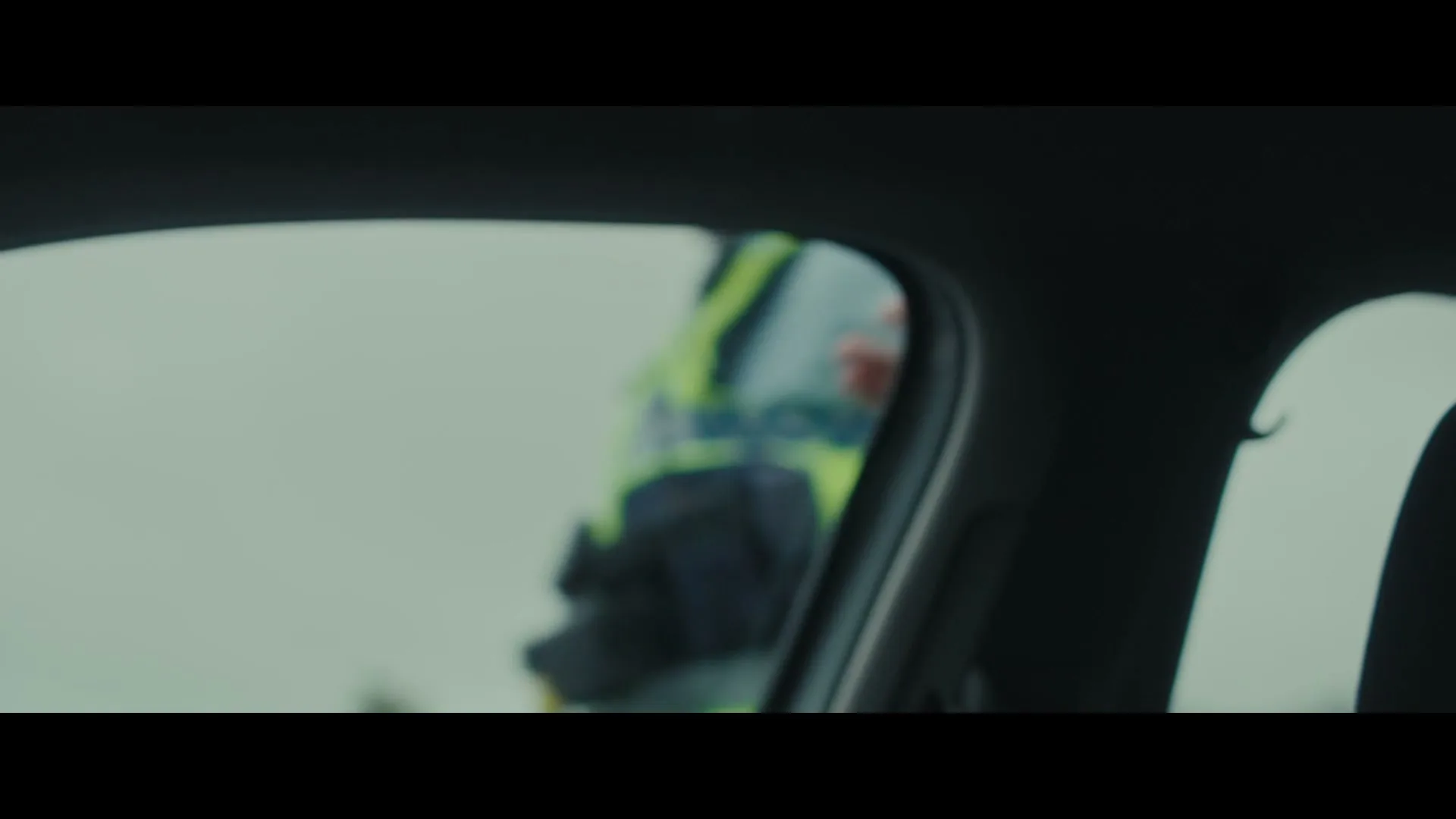 A blurry view from inside a car shows a person in a high-visibility vest standing outside, partially reflected in the side mirror.