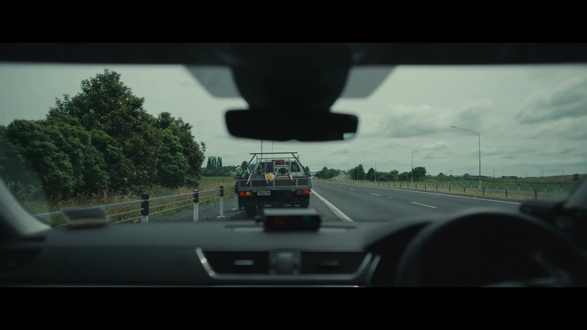 A POV shot from inside a car shows a utility truck with a worker in a high-vis vest on the side of a highway under a cloudy sky.