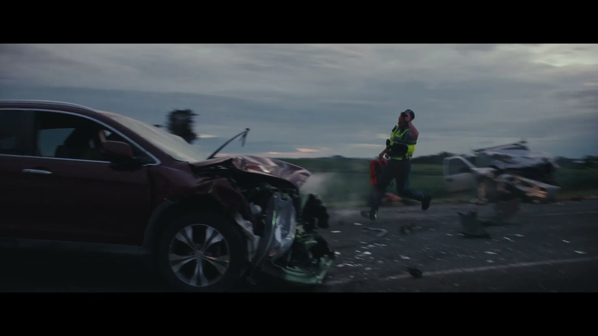 A man in a high-vis vest runs towards a crashed car on a rural road, with debris scattered around and other people in the background.