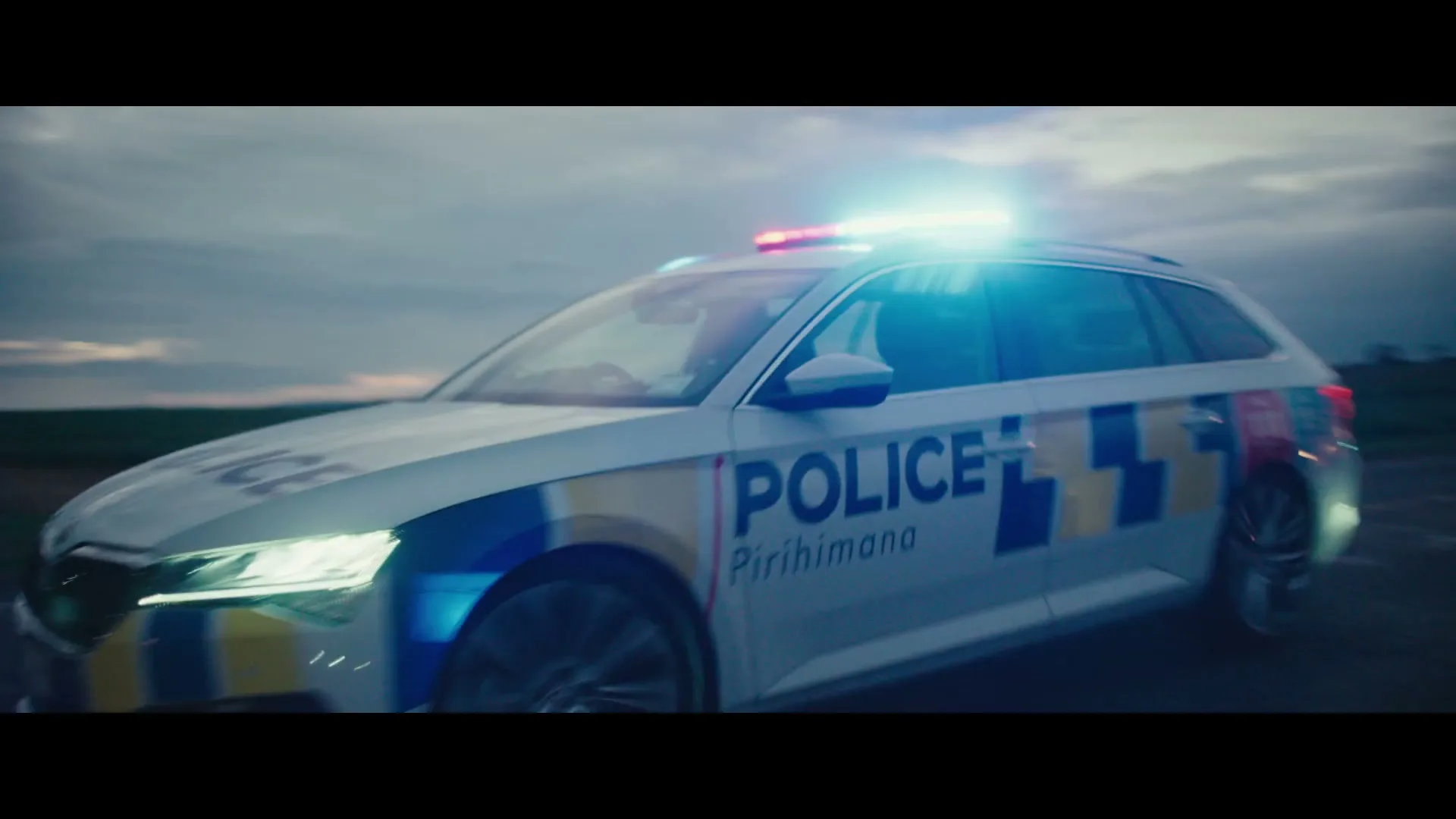 A white police car with flashing lights drives on a road at dusk, with a cloudy sky in the background.