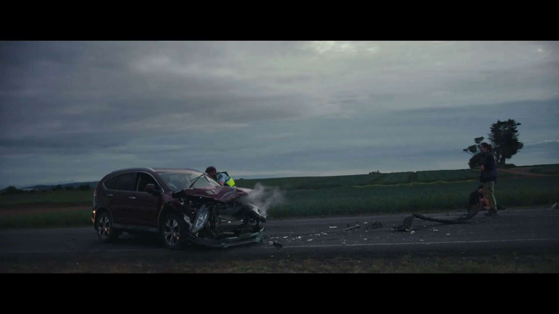 A crashed red SUV with a crumpled front end sits on the side of a rural road at dusk, with debris scattered around it and people standing nearby.