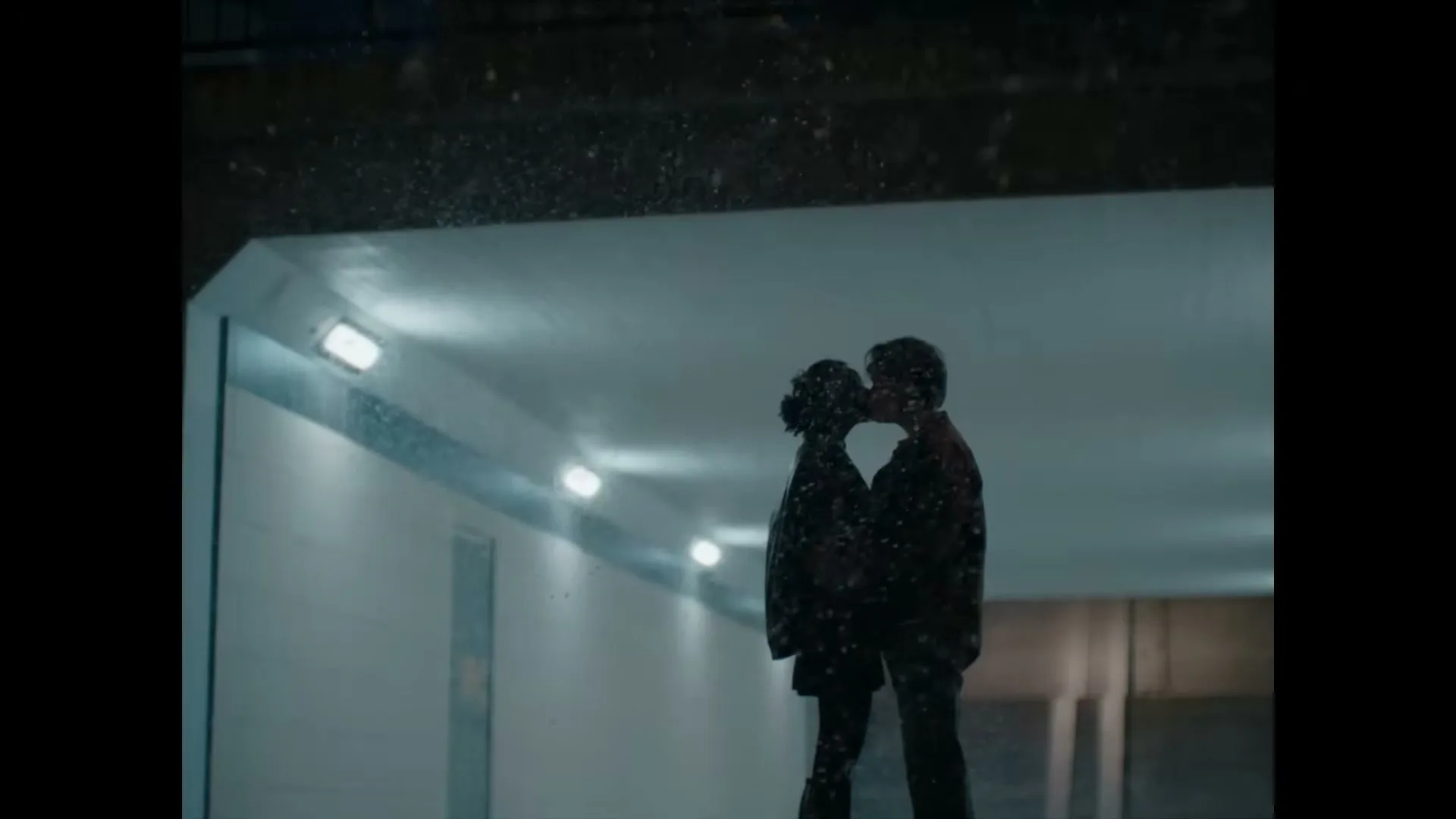 A young couple, silhouetted against a tunnel, kisses while snow falls around them at night.