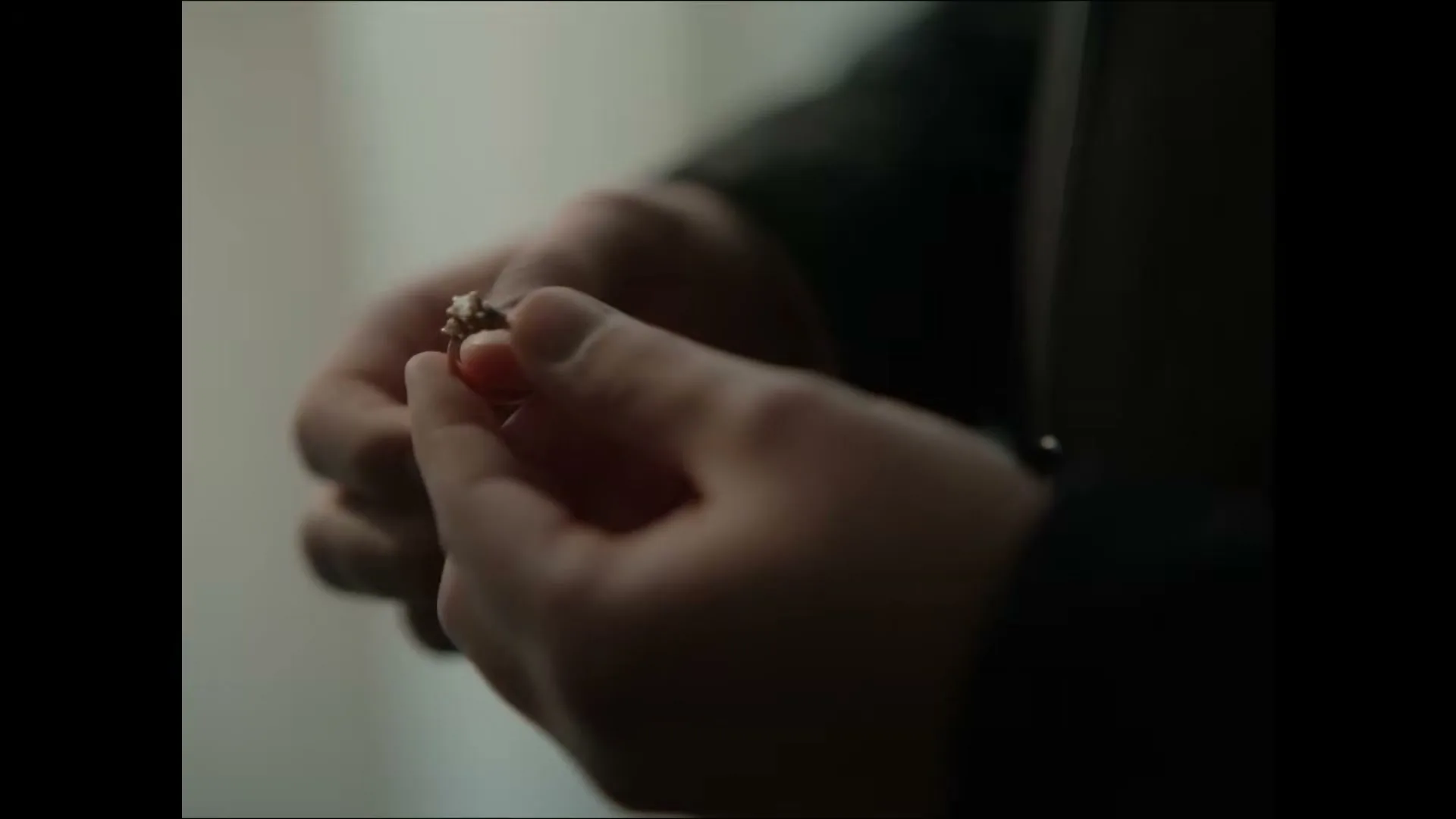 A man's hands hold a small, ornate ring, examining it closely in a dimly lit interior setting.