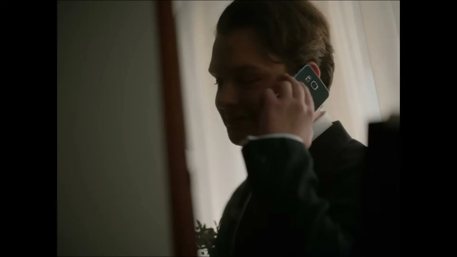 A young man in a suit holds a smartphone to his ear, smiling slightly, in a dimly lit interior.