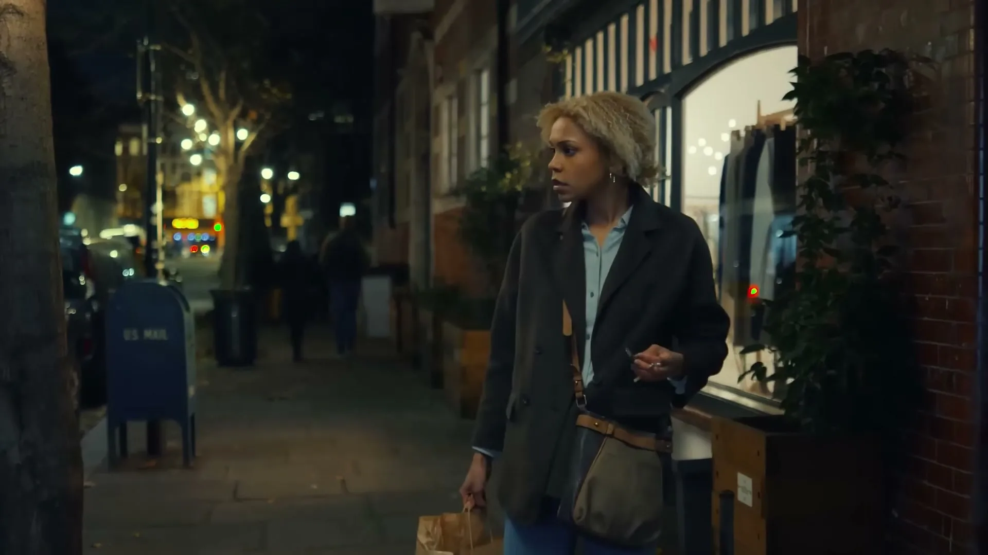 A young woman with blonde hair, wearing a dark coat and jeans, walks down a city street at night, looking around with a concerned expression.