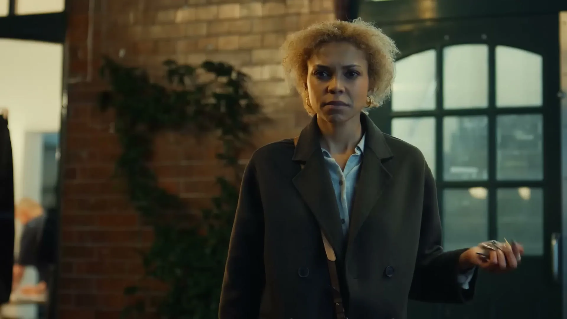 A woman with blonde curly hair, wearing a dark coat, stands holding keys, looking concerned in an interior setting.