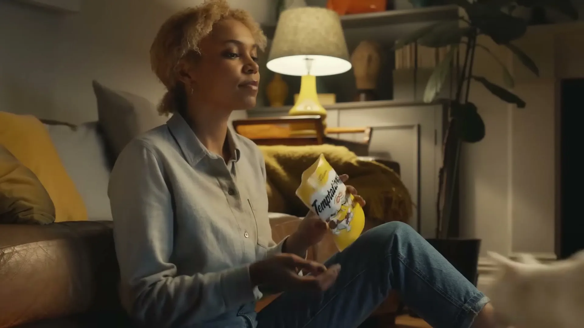 A young woman with curly blonde hair sits on the floor in a living room, holding a bag of Temptations cat treats as a white cat approaches her.
