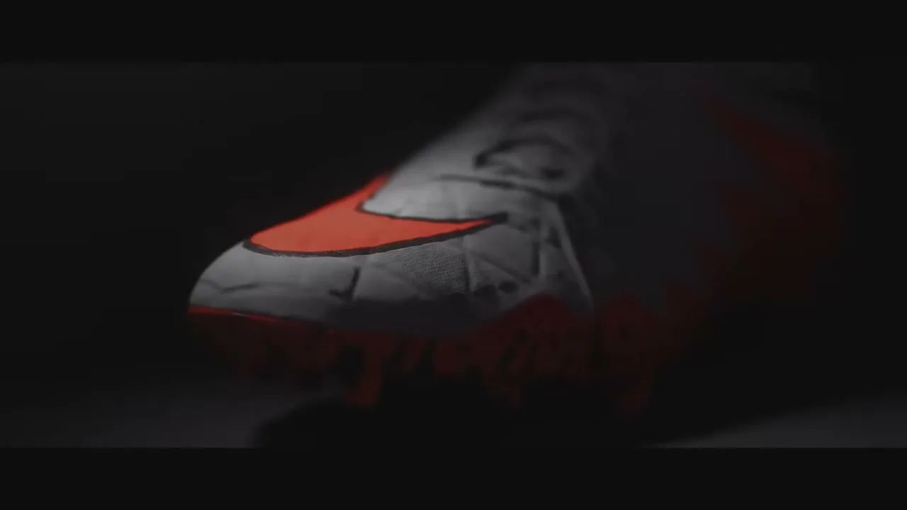 A white and orange Nike Hypervenom soccer cleat is shown in a dark setting, with a spotlight on the toe, then the cleat rotates to reveal the full shoe.