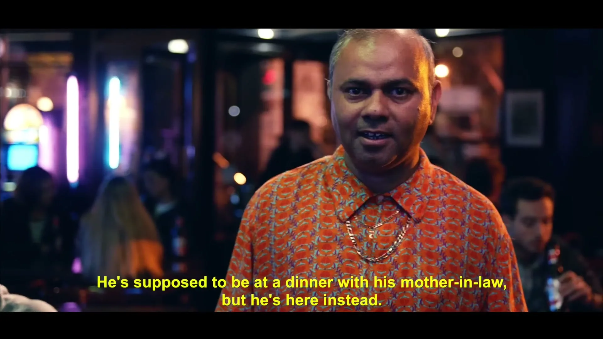 A man in an orange shirt and gold chain smiles at the camera, while two men at a bar table talk in the background.