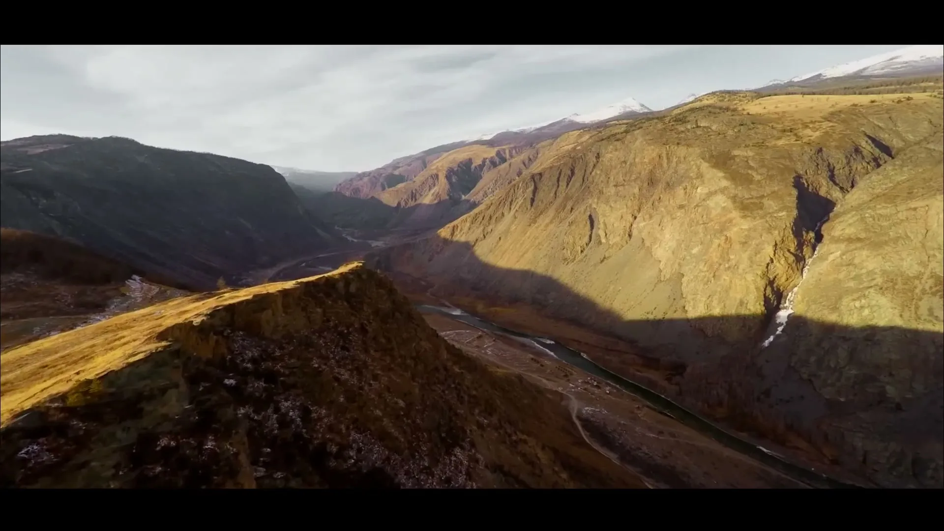 An aerial view captures a vast mountain valley with a river winding through it, under a bright sky with dramatic shadows.