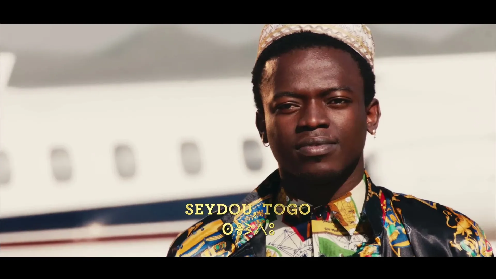 A young Black man in a patterned shirt and hat stands on an airport tarmac with a plane in the background, looking directly at the camera.