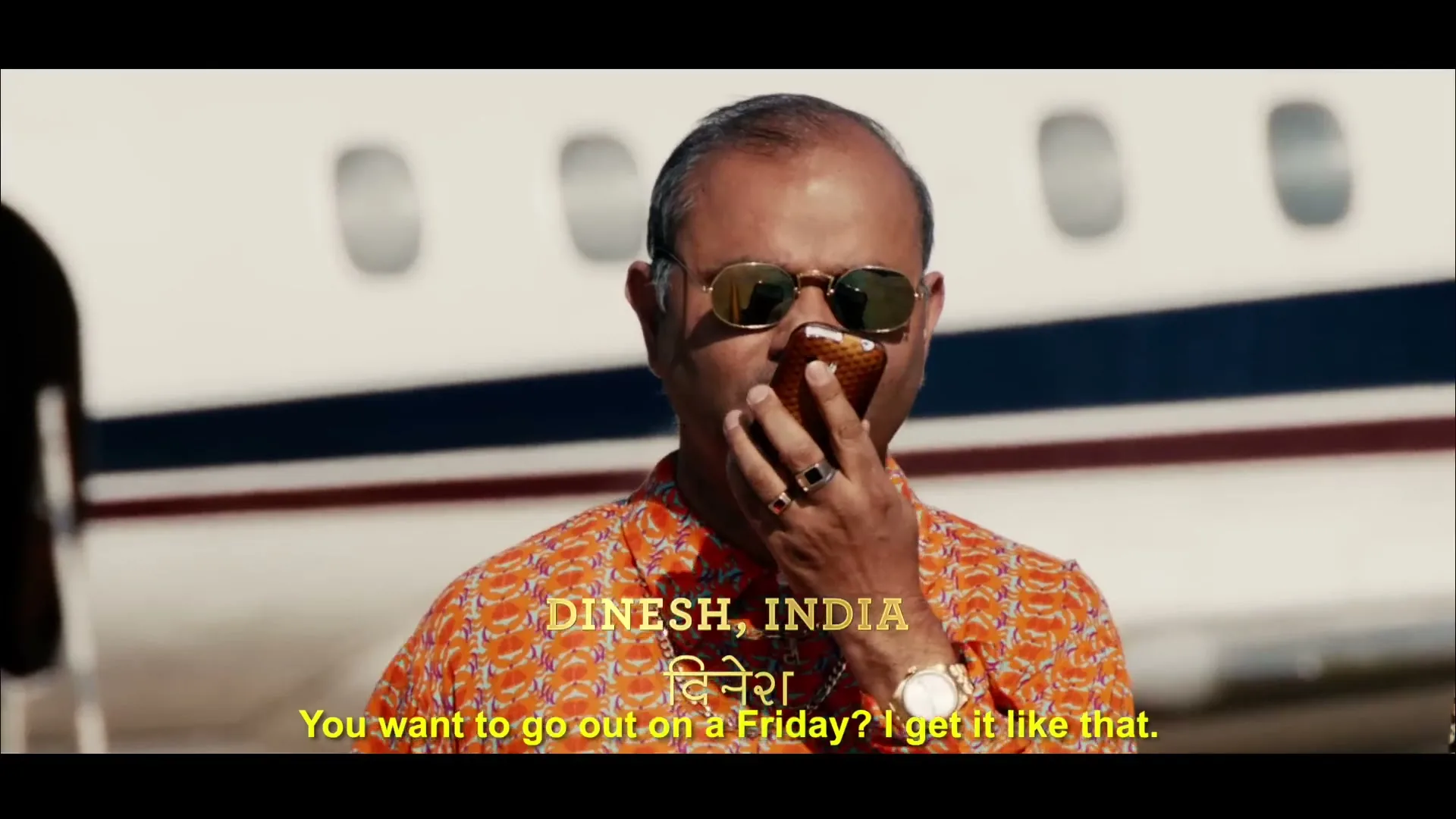 A man in sunglasses and a patterned shirt holds a phone to his mouth, speaking, with a private jet in the background on a sunny day.
