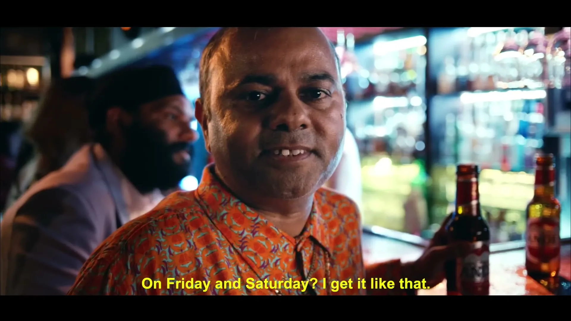 A man in his 40s, wearing an orange patterned shirt, talks to the camera while holding a beer bottle in a dimly lit bar.