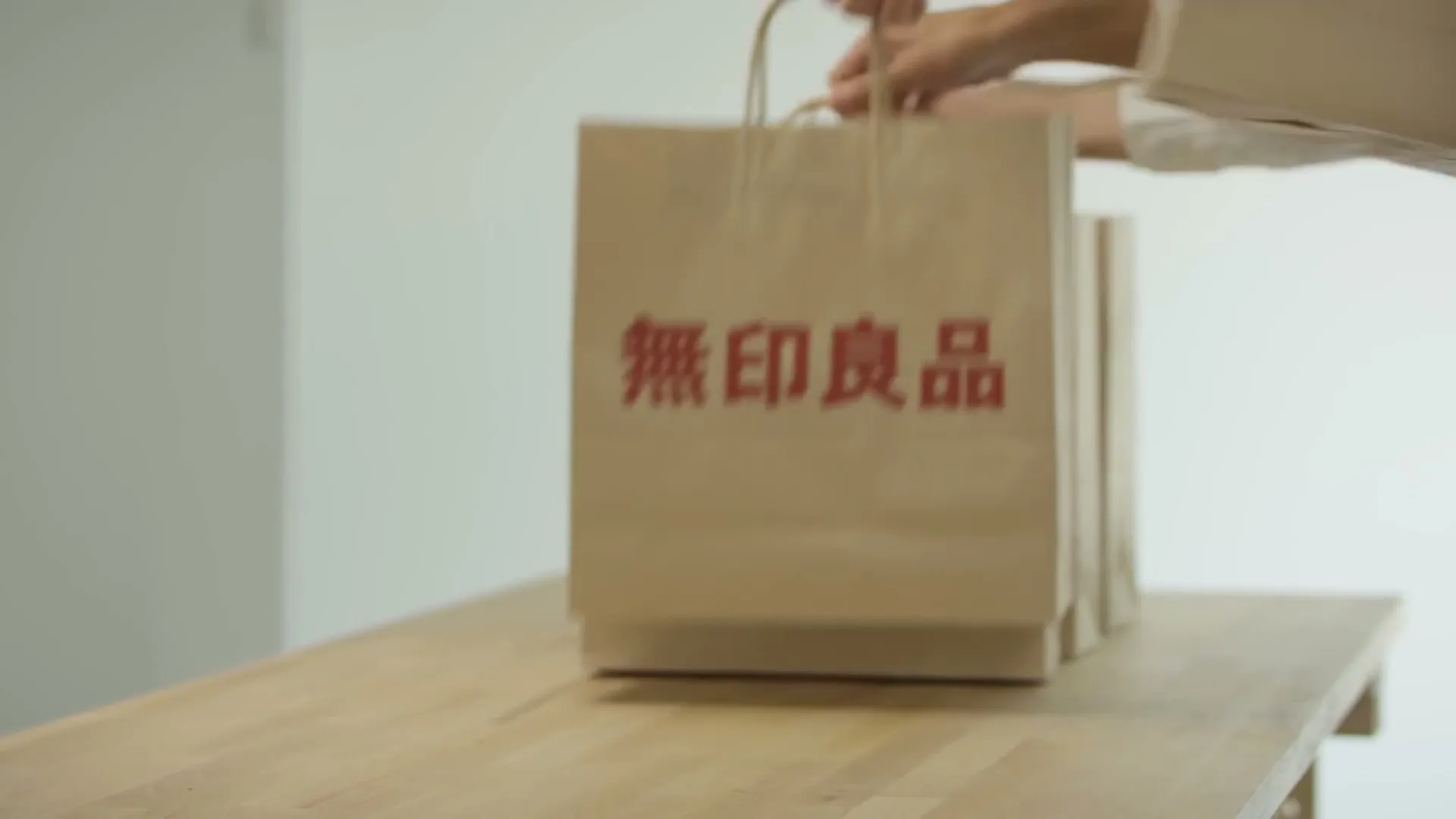 A person's hands place several brown paper shopping bags with red Japanese text onto a light wooden table in a brightly lit room.