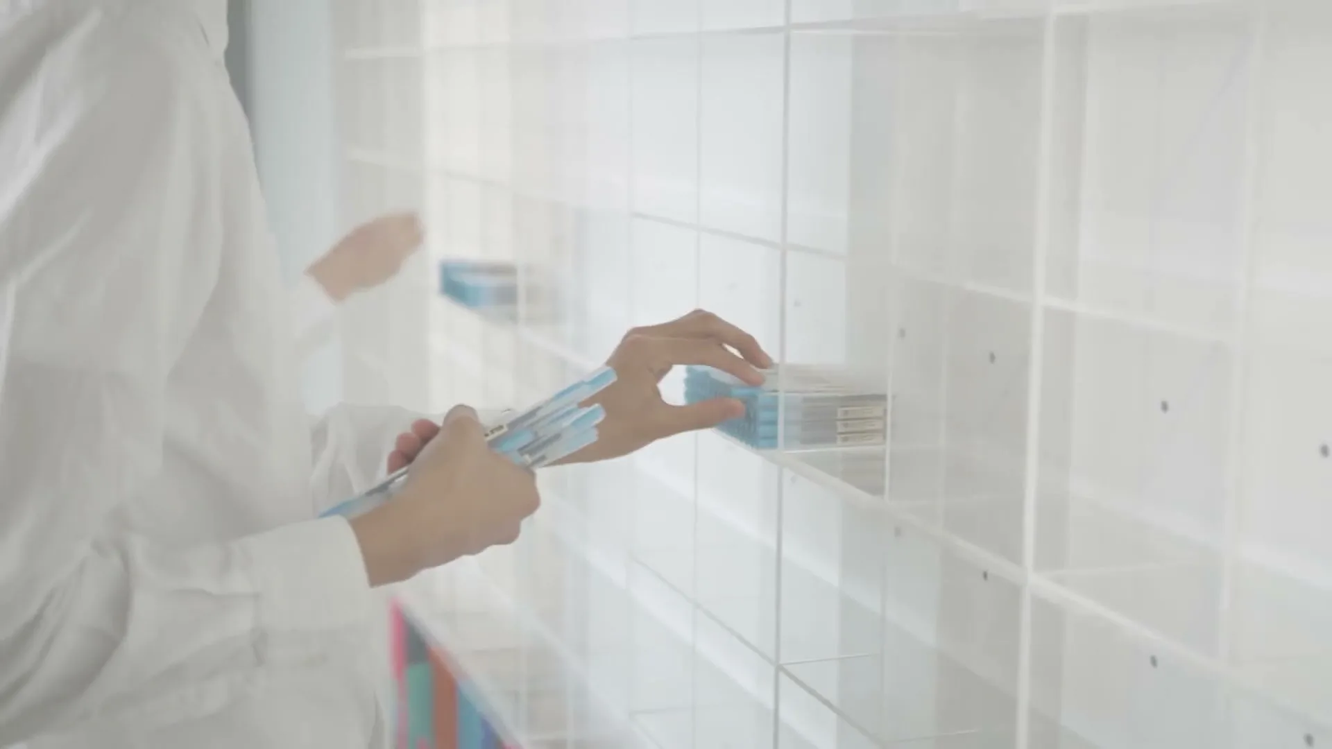 A person in a white lab coat places a blue pen into a clear, gridded shelf, holding several other pens.