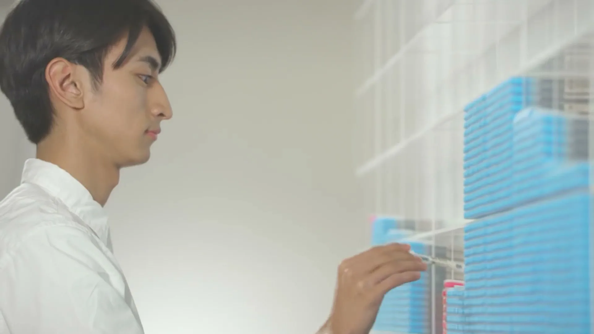 A young man in a white shirt uses a pen to interact with a grid of colorful blocks on a transparent surface in a bright, minimalist setting.