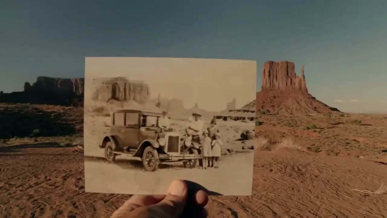A hand holds up an old black and white photo of a family and a vintage car in a desert landscape, matching the background.