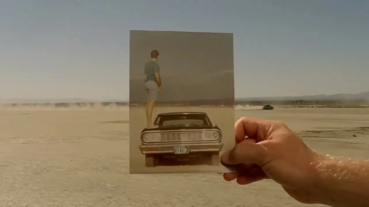 A man's hand holds up an old photograph of a young man standing next to a classic car in a desert landscape, aligning it with the current desert background.
