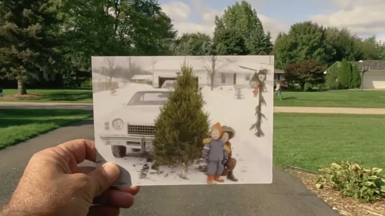 A hand holds up an old photograph of two children next to a Christmas tree and a car in a snowy suburban driveway, with a modern suburban street in the background.