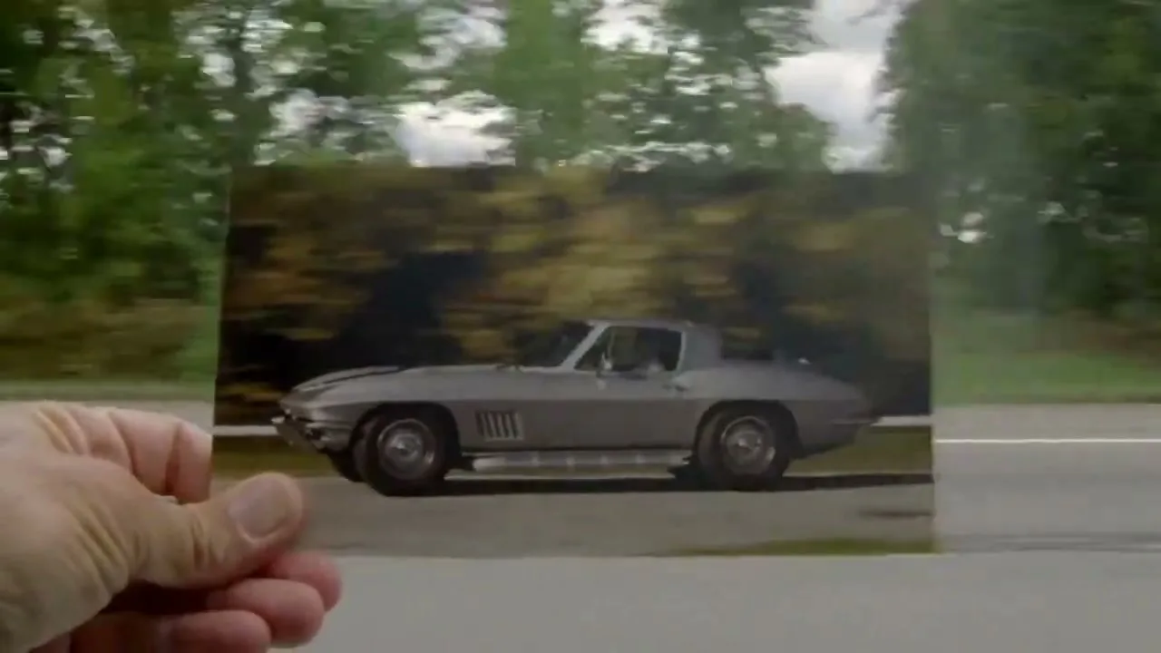 A person's hand holds up a photograph of a vintage silver Corvette, with a blurred background of trees and a road, suggesting movement.