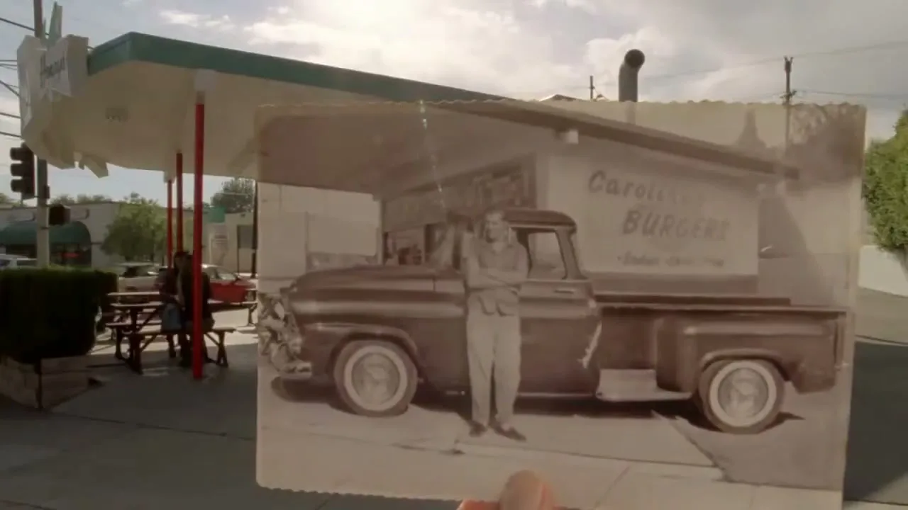 A hand holds up an old sepia photograph of a man standing next to a vintage Chevrolet pickup truck in front of a burger joint, aligning it with the present-day location.
