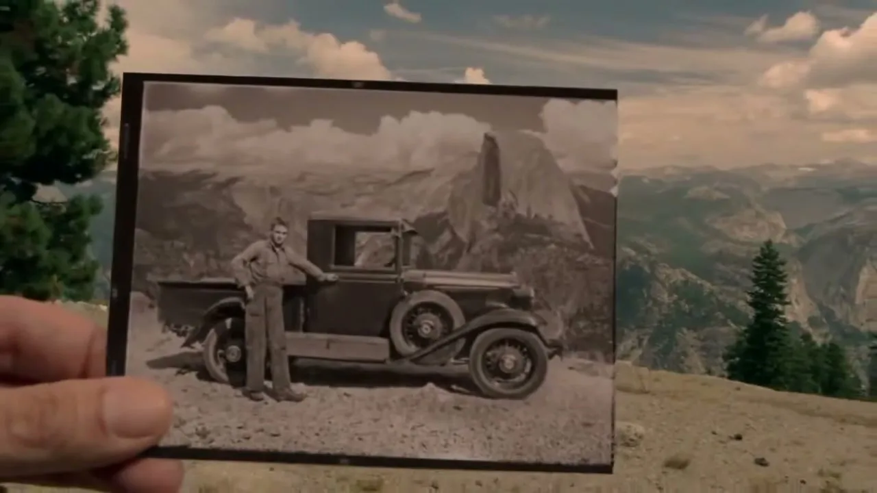 A hand holds up a black and white photo of a man and a vintage truck in front of Half Dome, Yosemite, matching the current landscape.