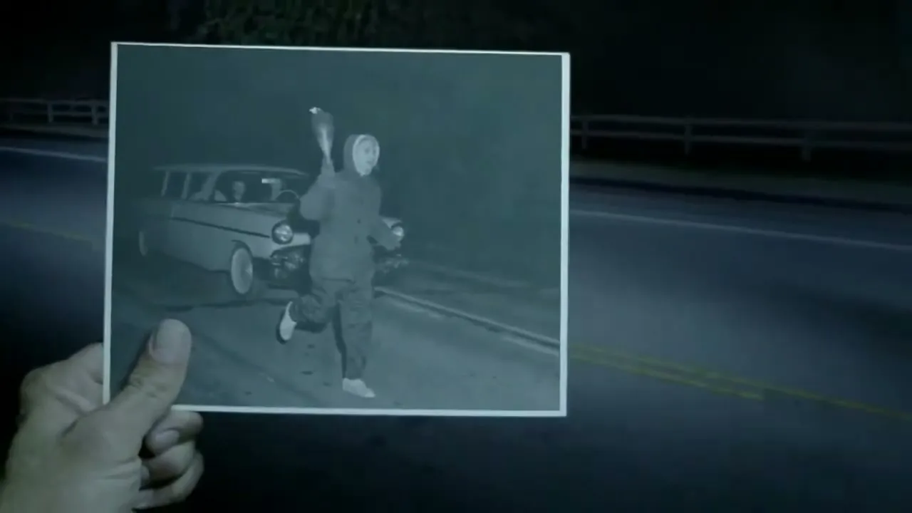 A hand holds up a black and white photo of a person running with a bottle, next to a vintage car on a dark road at night.