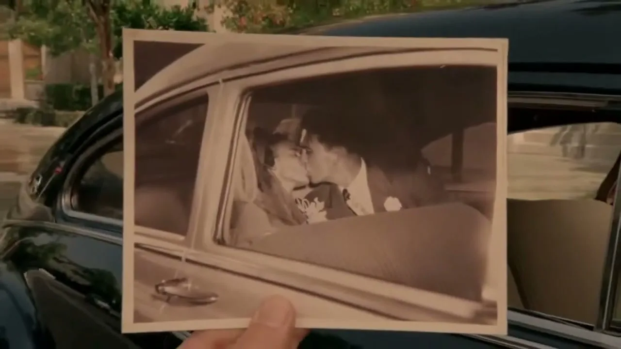 A hand holds up an old black and white photo of a newlywed couple kissing in a car, with a vintage car in the background.