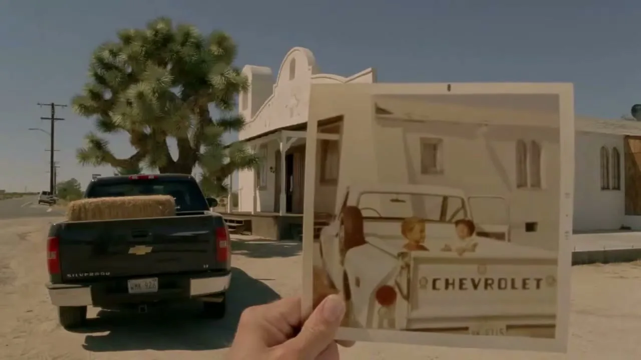 A hand holds up an old photograph of a white Chevrolet truck with two children in the bed, aligning it with a modern black Chevrolet truck and a desert building.