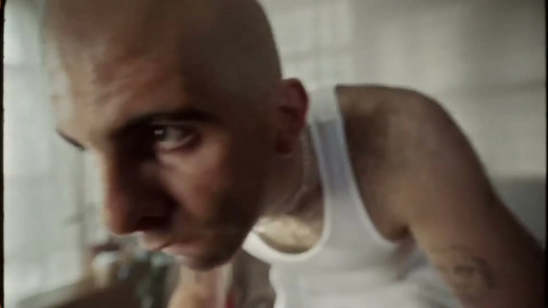 A bald man in a white tank top looks intently at something below him, with a focused expression in an interior setting.