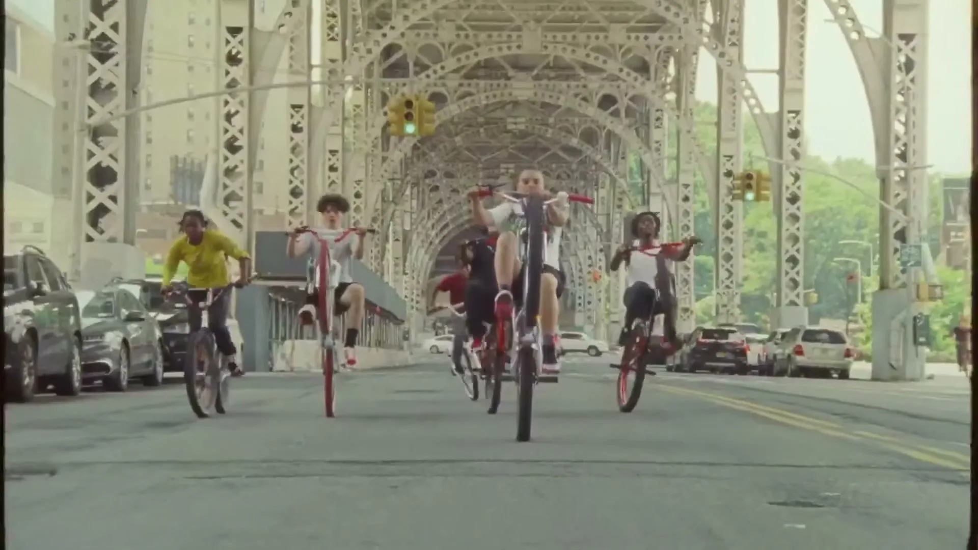 A group of young people ride bicycles on their back wheels down a city street under a large metal bridge.