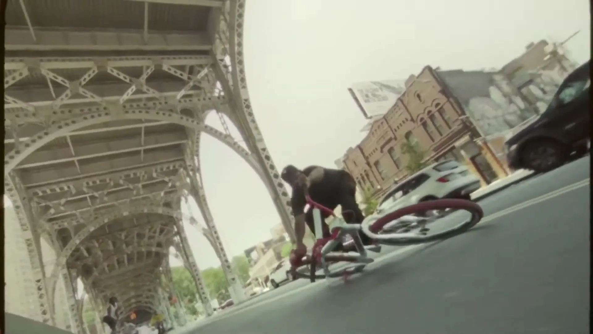 A male cyclist in a black shirt and pants performs a trick on his red bike under a large metal bridge on a city street.