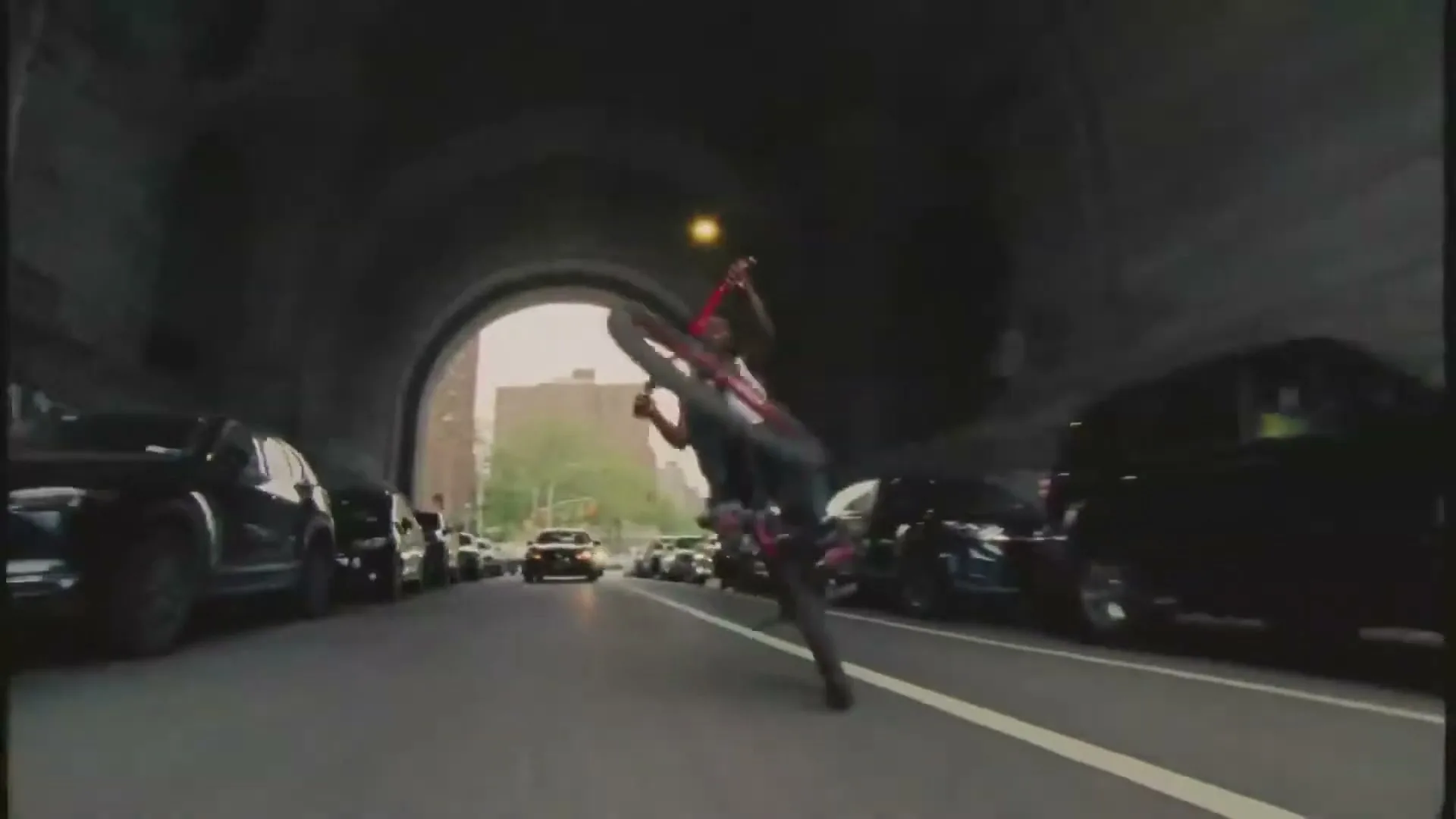A young man in a white tank top and dark pants performs a wheelie on a red bicycle, riding through a dark tunnel with parked cars on both sides.