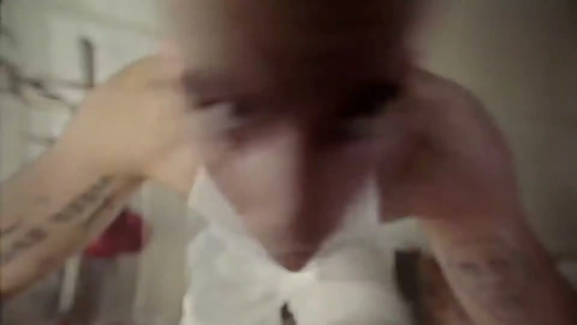 A young man with a shaved head and tattoos on his arms looks down at the camera, then a hand pushes a red toy into a black object.