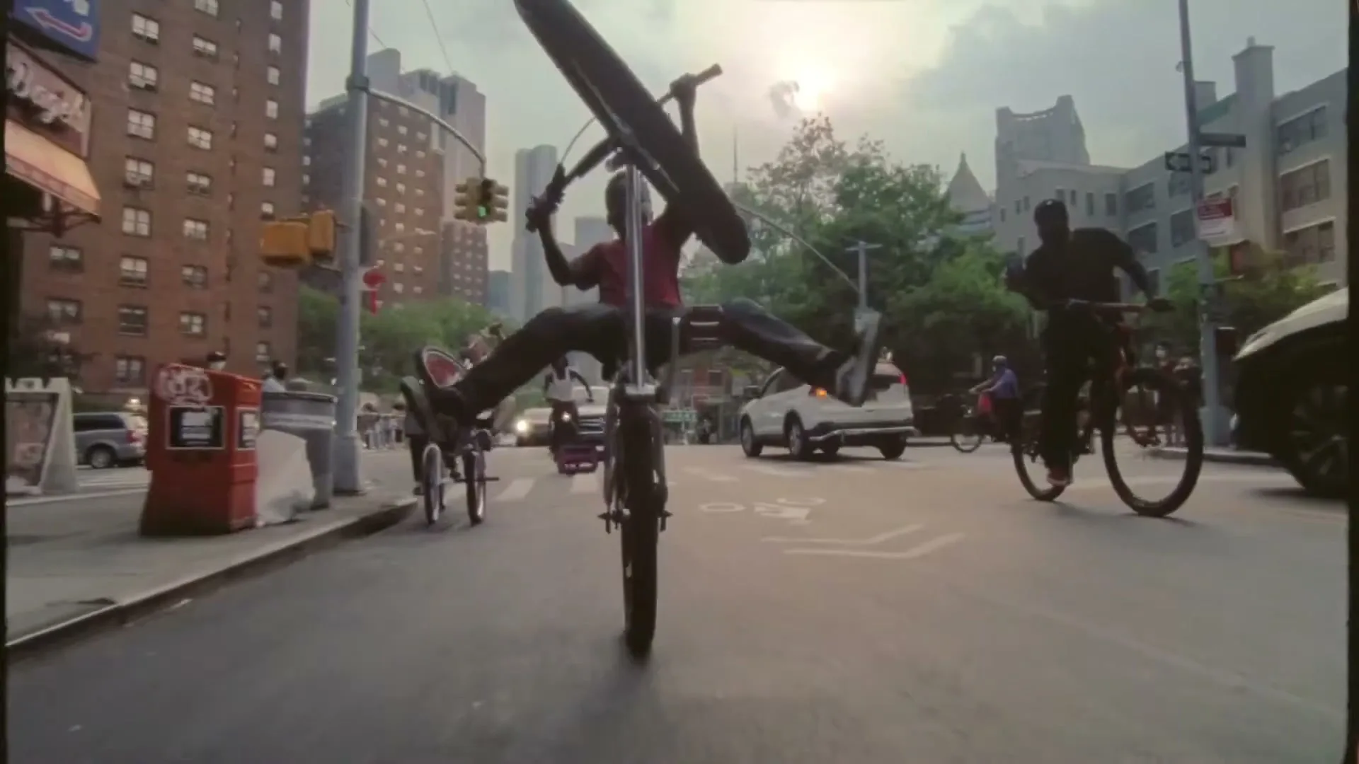 A young male biker in a red shirt and dark pants performs a wheelie on a city street, with other bikers and buildings in the background.