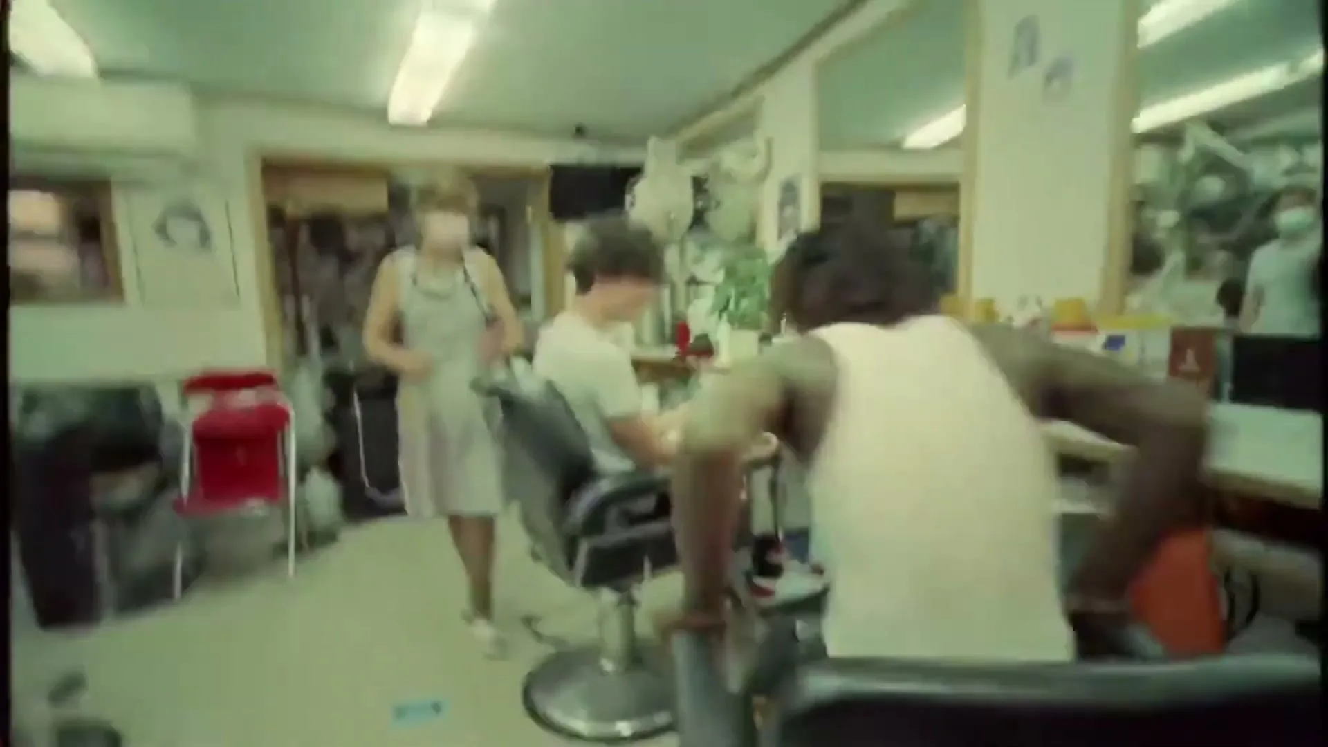 A young Black man in a white tank top and dark pants is seen from behind, leaning over a barber chair in a brightly lit barbershop.