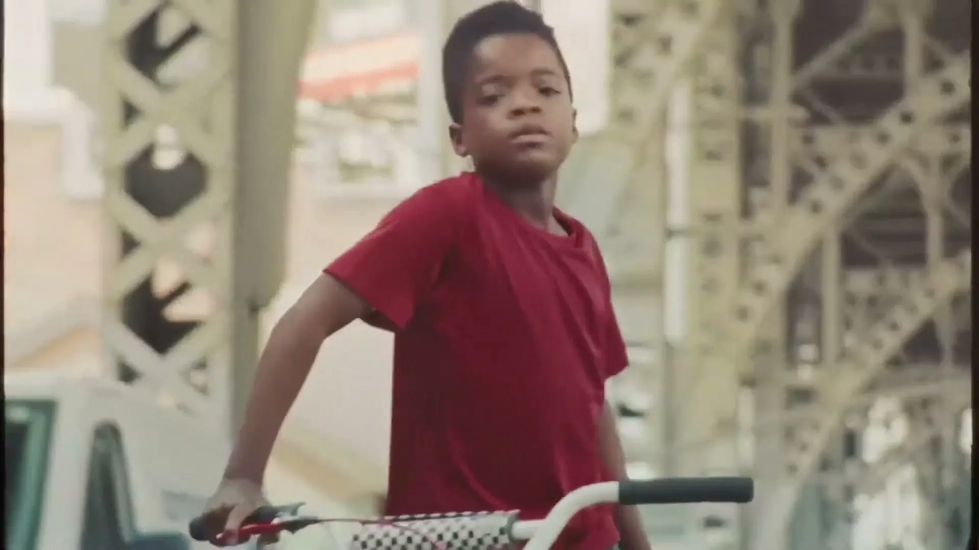 A young Black boy in a red t-shirt rides a BMX bike under a bridge, looking around with a determined expression.