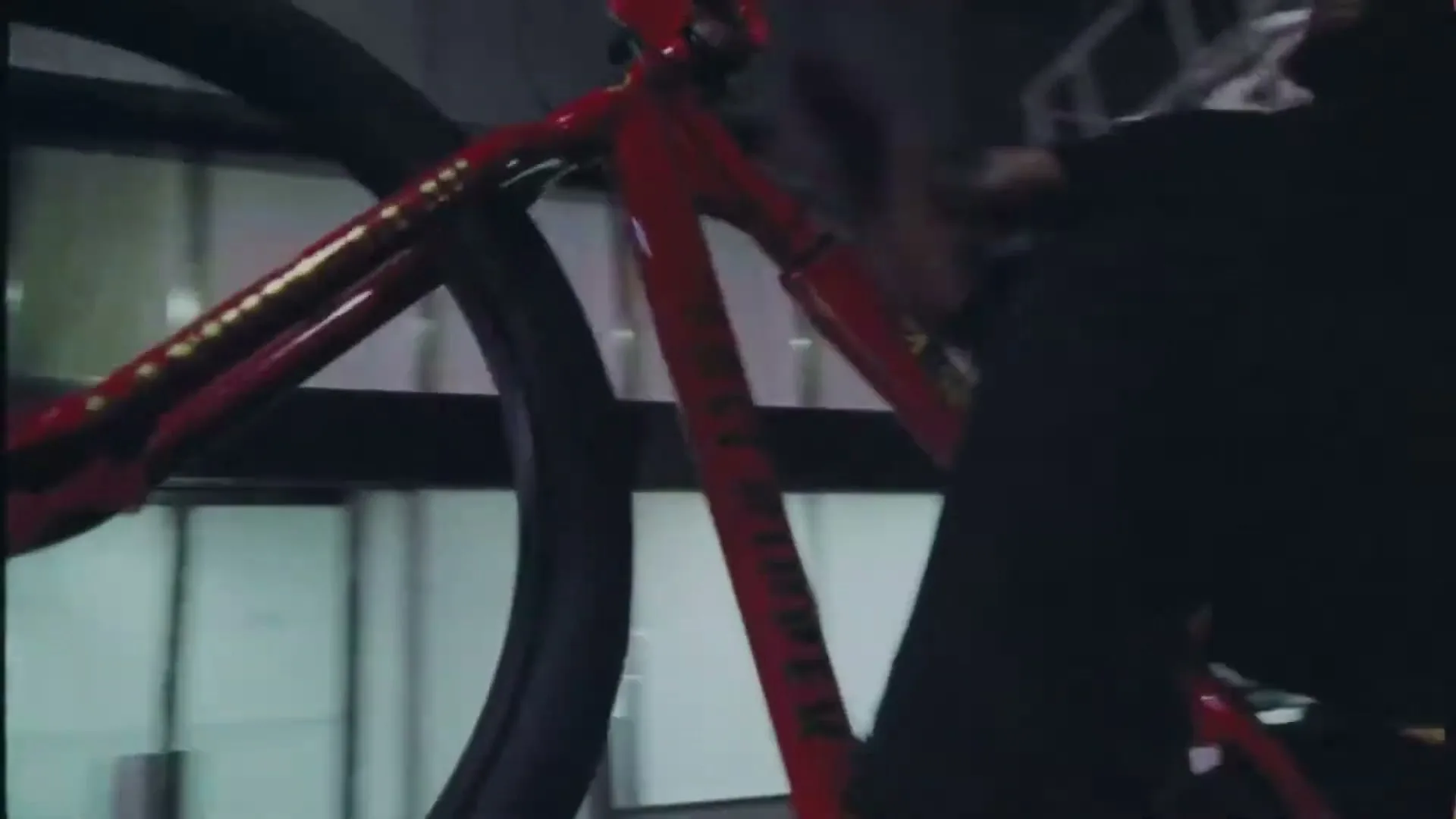 A person in a black shirt rides a red bicycle with gold accents, blurred against a background of buildings, suggesting fast movement.