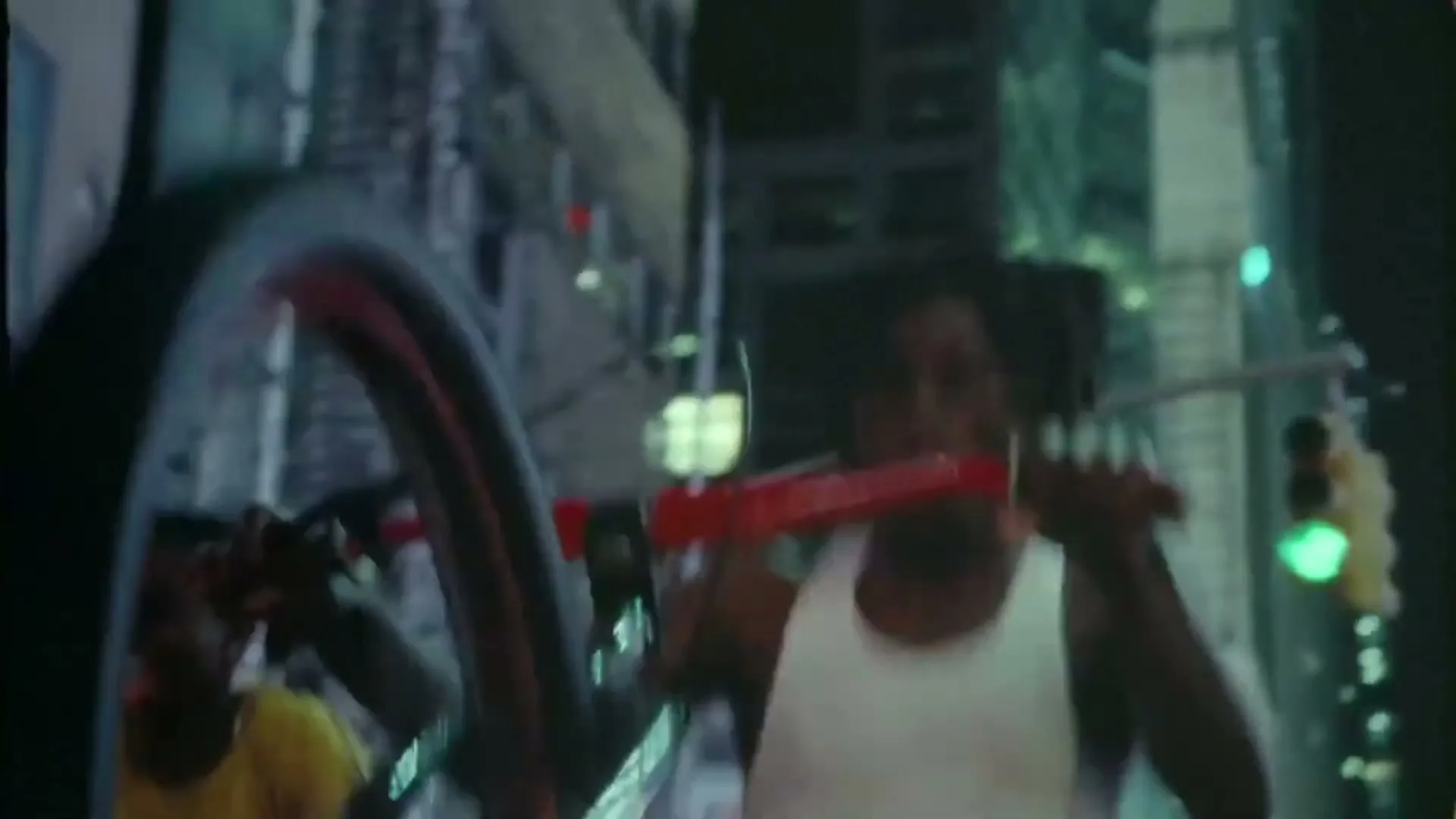 A young man with dreadlocks in a white tank top rides a red BMX bike at night in a city street, with another person visible behind him.