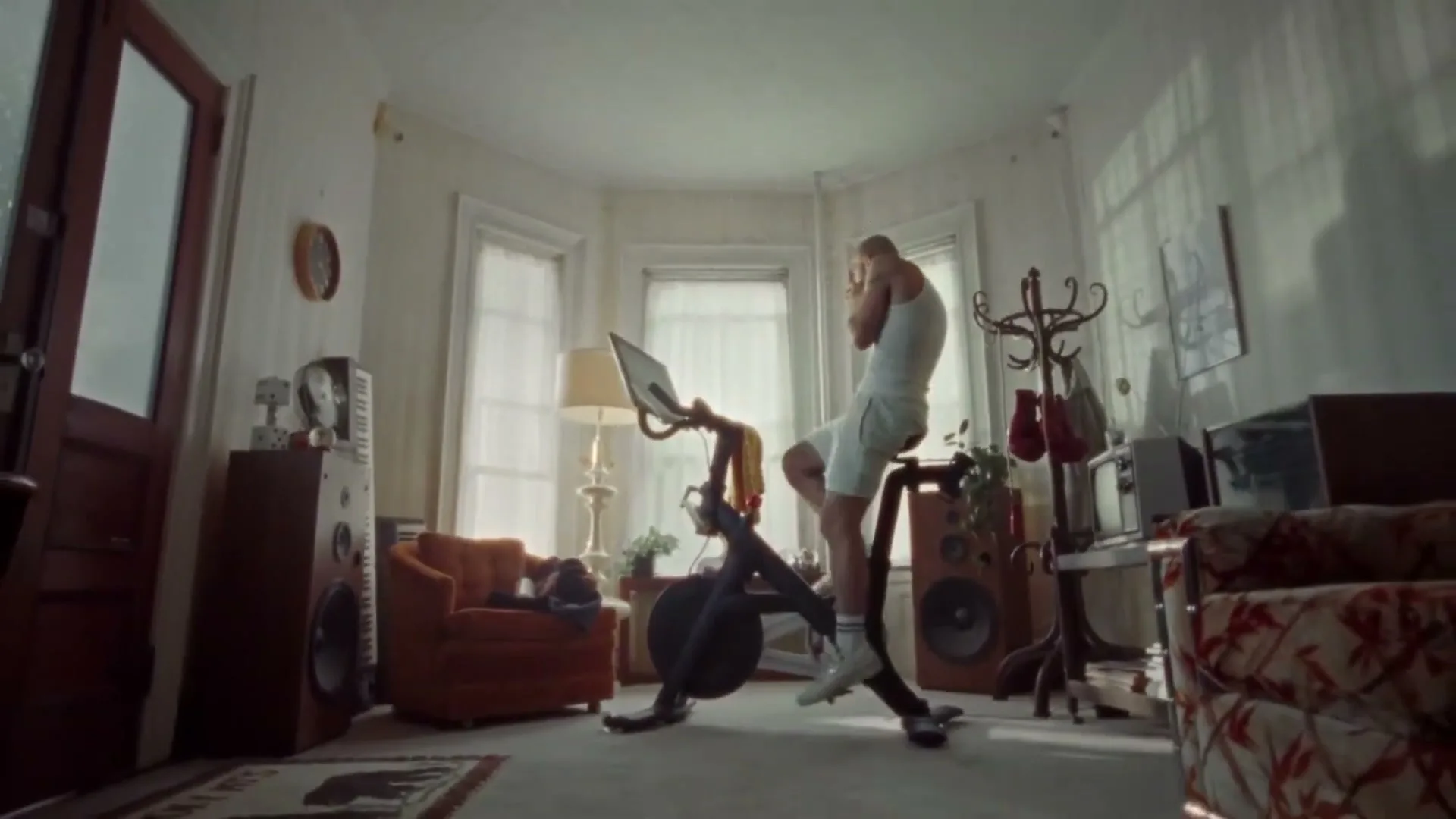 A bald man in his 30s, wearing a white tank top and shorts, rides a stationary bike in a cluttered living room, then slows down and stretches.