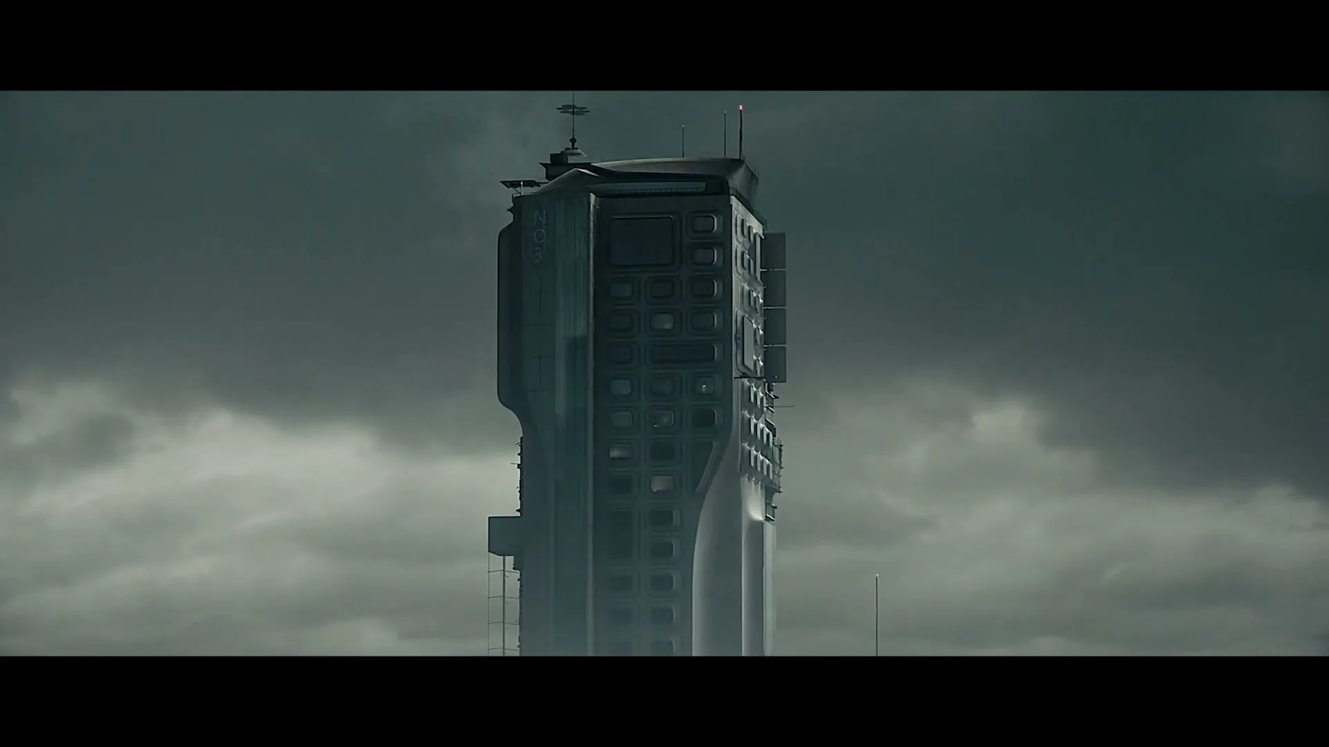 A tall, futuristic building stands amidst a cloudy, grey sky, with a single window on its facade glowing brightly.