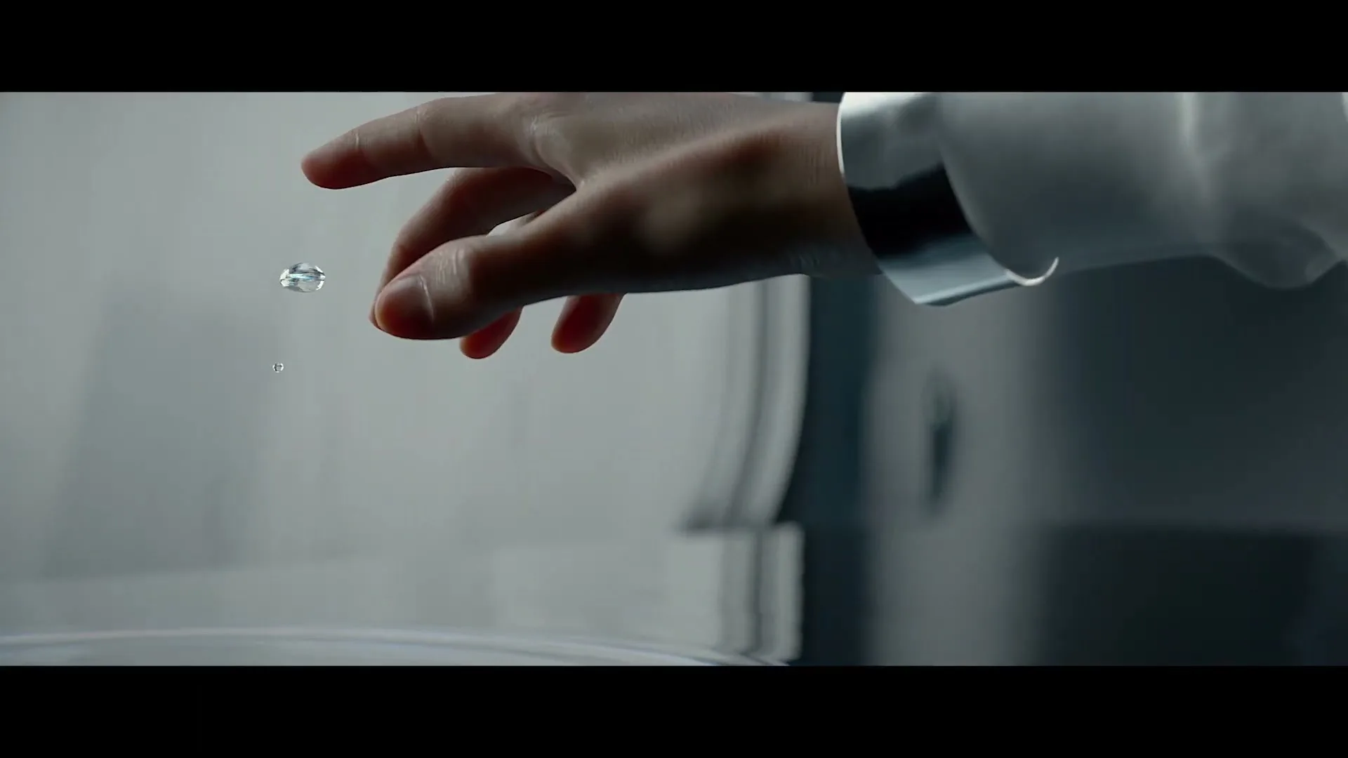 A person's hand in a white sleeve reaches out as a small droplet of water floats in the air above a water surface.